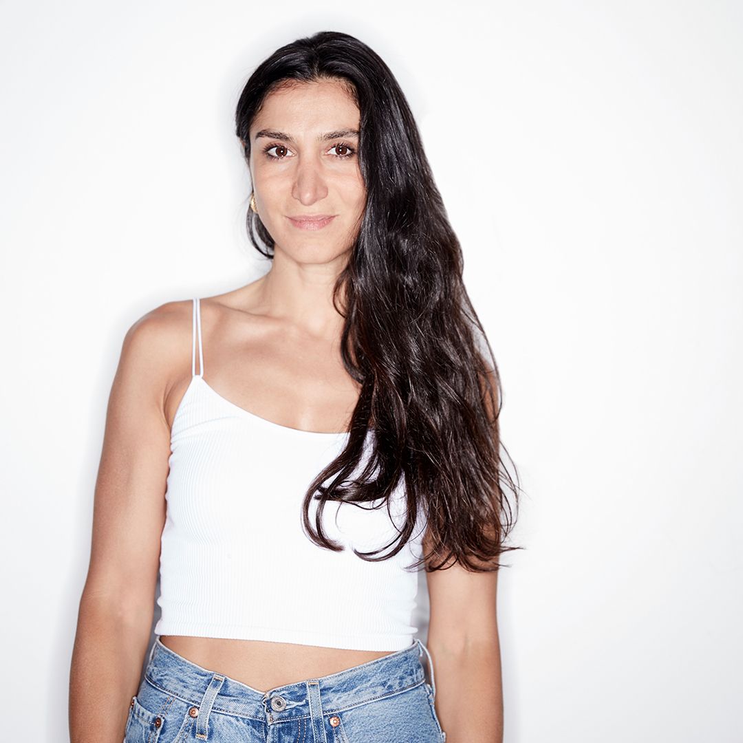 image of women with long dark hair standing looking into the camera, wearing a white shirt and jeans
